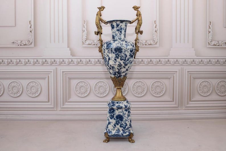 May include: A large, blue and white porcelain vase with gold accents and a decorative base. The vase is decorated with a floral pattern and features two gold figures on the handles.
