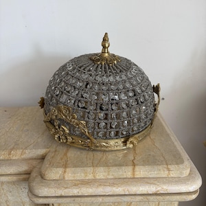 May include: A dome-shaped ceiling light fitting featuring a gold-toned metal frame with ornate floral detailing at the base and a central finial. The structure is covered in a mesh of small grey beads interspersed with clear, faceted glass crystals.