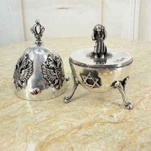 May include: A silver-toned metal container with a removable dome-shaped lid. The lid features a crown finial and double-headed eagle emblems. The base has three legs, a Star of David motif, and a small figurine handle on its flat cover.