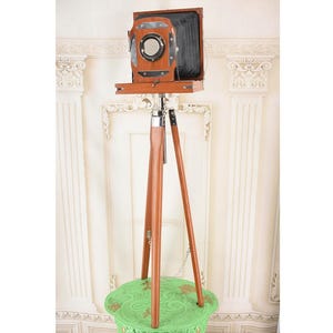 May include: A vintage wooden camera with a large lens and bellows, mounted on a wooden tripod. The camera is brown with black accents and a silver metal lens ring. The tripod has three legs and is also made of wood. The camera is positioned on a green table.