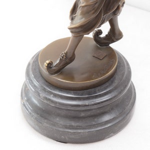 Op de afbeelding: Bronzen beeldje van een persoon in een vloeiende jurk en puntige schoenen, staande op een zwarte marmeren voet. Het beeldje is gesigneerd op de voet.