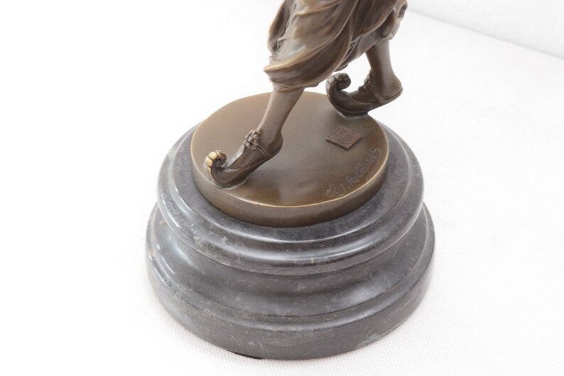 Op de afbeelding: Bronzen beeldje van een persoon in een vloeiende jurk en puntige schoenen, staande op een zwarte marmeren voet. Het beeldje is gesigneerd op de voet.