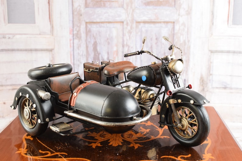 May include: A detailed black and brown motorcycle with a sidecar. The motorcycle features a brown leather seat, a small suitcase, and a spare tire on the sidecar. The wheels have a gold finish, and the headlights are a bright gold color.