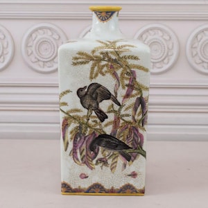 Op de afbeelding: Rechthoekige keramische vaas met een crèmekleurige achtergrond, met illustraties van vogels en botanische elementen in bruin, paars en groen. De vaas heeft een gele rand en basis met decoratieve patronen. Hoogte ongeveer 25 cm.
