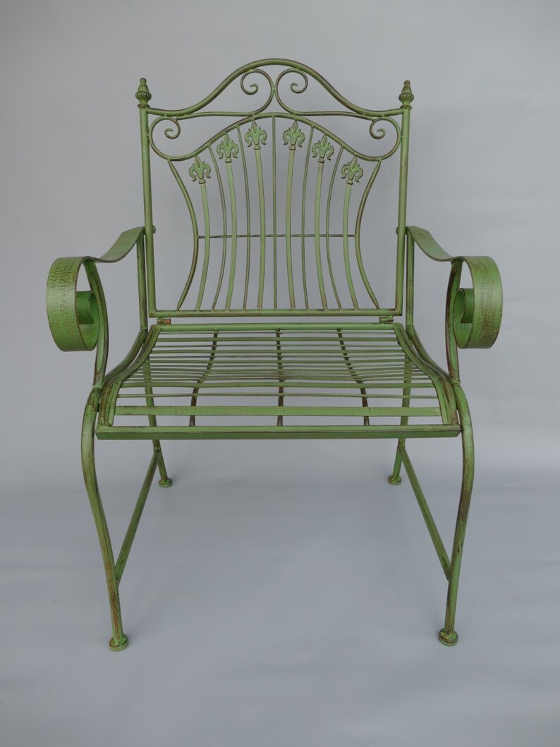 May include: A green metal garden chair with a decorative back featuring fleur-de-lis accents. The chair has curved armrests and a slatted seat. The overall design is ornate and vintage, with a distressed finish.
