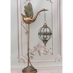 May include: A bronze metal floor lamp with a swan design. The swan holds a glass globe lantern. The lamp has a decorative base and a curved support arm. The background is a light-coloured wall with ornate moulding. The lamp is approximately 150 cm tall.