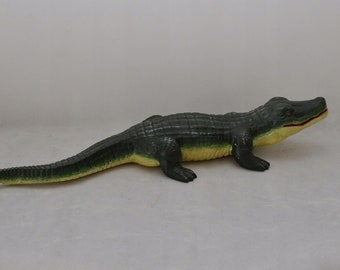 May include: A green and yellow plastic alligator toy. The alligator is lying on its belly with its head turned to the right. The toy has a textured surface to resemble scales.