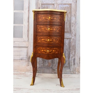 May include: A small, antique wooden cabinet with four drawers. The cabinet is a dark brown with decorative inlaid patterns and gold accents. The legs are curved, and the top is trimmed with gold.