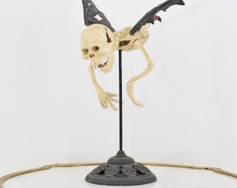 May include: A skeletal figure with bat wings, a skull head, and a fanged mouth is mounted on a black stand. The figure is posed in mid-flight with its wings spread wide.