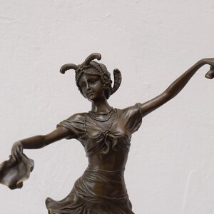Op de afbeelding: Bronzen beeld van een vrouw in een vloeiende jurk, die in elke hand een stuk stof vasthoudt. Ze draagt een hoofdtooi met veren en een ketting. Het beeld is in klassieke stijl.