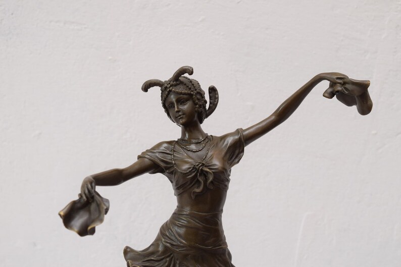 Op de afbeelding: Bronzen beeld van een vrouw in een vloeiende jurk, die in elke hand een stuk stof vasthoudt. Ze draagt een hoofdtooi met veren en een ketting. Het beeld is in klassieke stijl.
