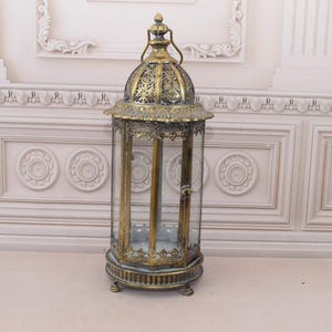May include: A decorative brass-effect metal lantern with a glass enclosure. The lantern features an ornate, domed top and a detailed base. The glass panels allow light to pass through. Suitable for candles or decorative items. The height is approximately 40 cm.