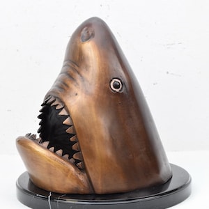 May include: Bronze sculpture of a shark head with an open mouth and sharp teeth. The sculpture is mounted on a black marble base.