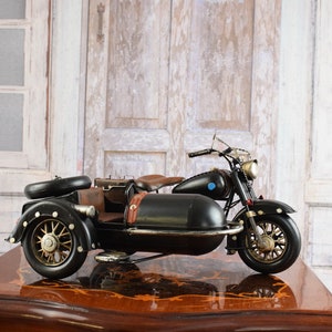 May include: A black motorcycle with a sidecar, featuring gold-colored wheels and accents. The motorcycle has a brown leather seat and luggage. The model sits on a wooden table with a decorative pattern. The background includes a white window and a wooden door.