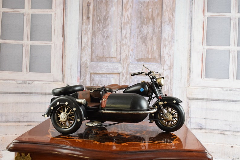 May include: A black motorcycle with a sidecar, featuring gold-colored wheels and accents. The motorcycle has a brown leather seat and luggage. The model sits on a wooden table with a decorative pattern. The background includes a white window and a wooden door.