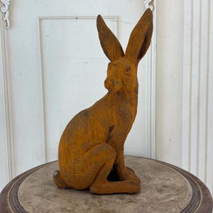 May include: A rust-colored, textured hare figurine in a seated position with long, upright ears. The sculpture features a weathered, oxidized metal appearance with visible fur-like detailing across its body and a small, rounded tail.