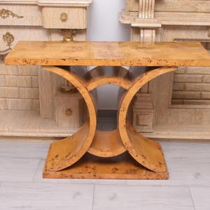 May include: A light brown, Art Deco style console table. The table has a rectangular top and a base with curved supports. The wood has a speckled pattern. The table is approximately 76 cm long.