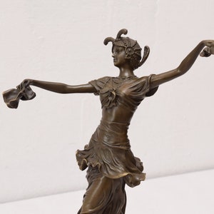 Op de afbeelding: Bronzen beeld van een vrouw in een vloeiende jurk, dansend met haar armen uitgestrekt, met stof in elke hand. Ze draagt een hoofdtooi met veren.