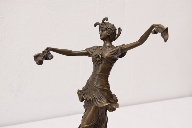 Op de afbeelding: Bronzen beeld van een vrouw in een vloeiende jurk, dansend met haar armen uitgestrekt, met stof in elke hand. Ze draagt een hoofdtooi met veren.