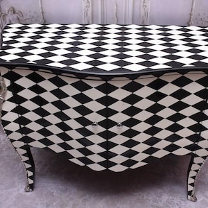 Puede incluir: Cómoda con estampado de arlequín en blanco y negro. La cómoda tiene una parte superior y patas curvas, con detalles decorativos plateados. El patrón de diamantes cubre toda la superficie de la cómoda.