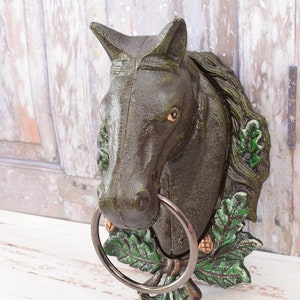 Op de afbeelding: Een handdoekring van gietijzer in de vorm van een paardenkop met groene bladeren en eikels. De ring is van metaal en is bevestigd aan het paardenhoofd.