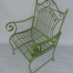 May include: A green metal garden chair with a scrolled back and armrests. The chair features a slatted seat and back, with decorative fleur-de-lis accents. The chair's dimensions are approximately 36 inches high, 24 inches wide, and 24 inches deep.