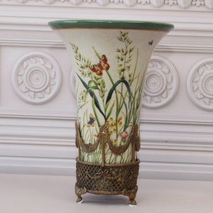 May include: A decorative vase with a cream-colored background, featuring a floral and butterfly design. The vase has a green rim and is supported by a bronze-colored metal base with ornate detailing. The vase is approximately 12 inches tall.
