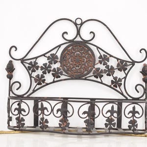 May include: Ornate, dark brown metal wall shelf with a decorative design. The shelf features a curved top with floral and scrollwork details, a central medallion, and a lower shelf for storage. The metal has a distressed finish.
