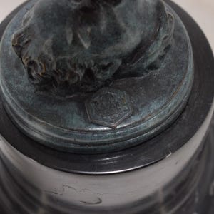 May include: A dark bronze sculpture of a classical bust on a black marble base. The bust has detailed facial features and is mounted on a round, tiered base. A small, hexagonal emblem is visible.