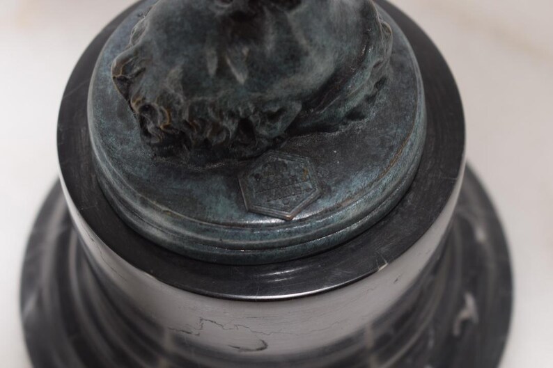 May include: A dark bronze sculpture of a classical bust on a black marble base. The bust has detailed facial features and is mounted on a round, tiered base. A small, hexagonal emblem is visible.