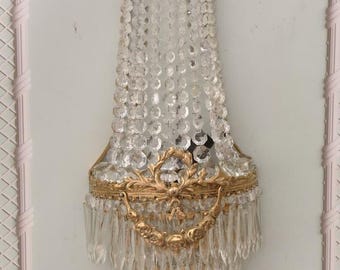May include: An ornate wall sconce with a gold-colored metal frame and clear crystal accents. The sconce features a cascading design of crystal beads and icicle-shaped drops, with decorative gold flourishes.