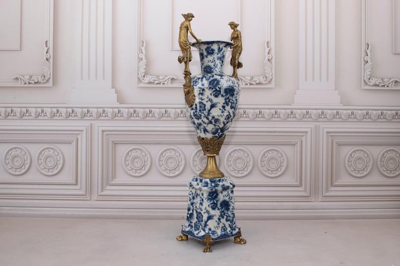 May include: A large, blue and white porcelain vase with gold accents and two gold cherub figures on the sides. The vase is on a pedestal with a matching blue and white design.