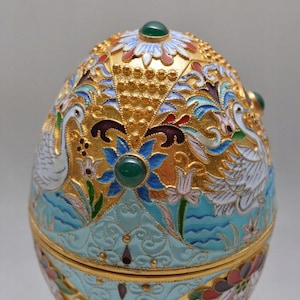 May include: A decorative egg-shaped trinket box with a blue and white enamel design featuring swans and flowers. The box is adorned with gold accents and green gemstones.