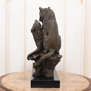 May include: Bronze sculpture of two wolves. One wolf is seated, the other is howling. The sculpture is mounted on a black rectangular base. The wolves are detailed with textured fur and are posed on a rocky outcropping.