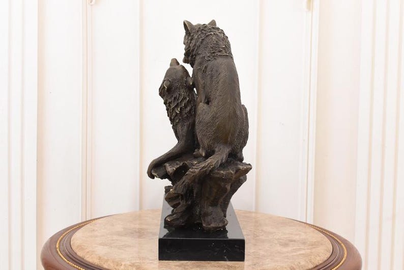 May include: Bronze sculpture of two wolves. One wolf is seated, the other is howling. The sculpture is mounted on a black rectangular base. The wolves are detailed with textured fur and are posed on a rocky outcropping.