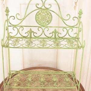 May include: A light green, ornate metal shelf with two tiers and decorative scrollwork. The shelf has a circular design in the center and is supported by slender legs. The shelf is placed on a round table with a marble-like top.