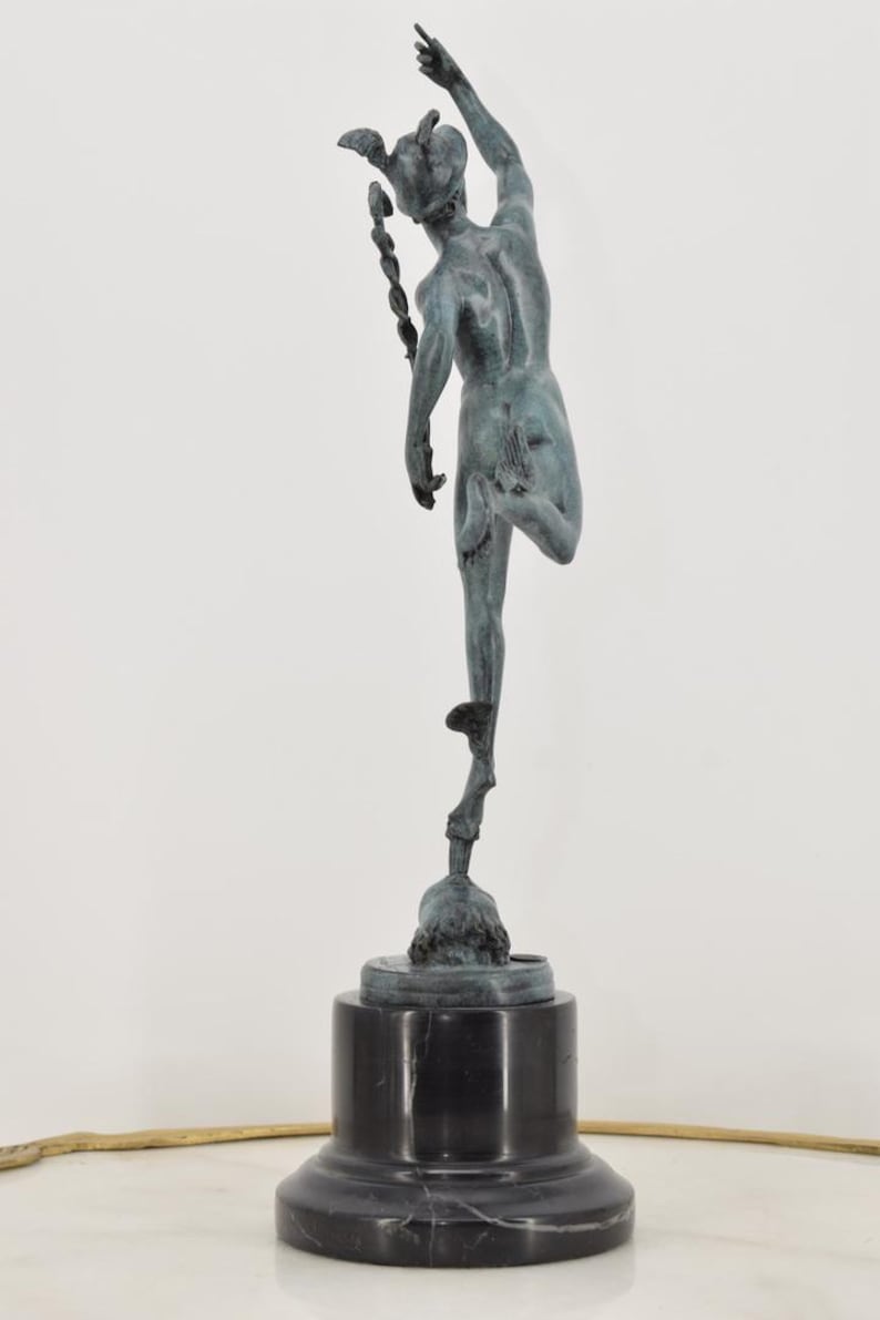 May include: A bronze sculpture of a figure in motion, poised on one leg atop a pedestal. The figure is reaching upwards with one arm, and has a winged helmet. The sculpture is mounted on a black marble base.