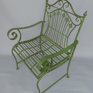 May include: A green metal garden chair with a decorative back featuring fleur-de-lis accents. The chair has curved armrests with scroll details and a slatted seat. The overall dimensions are approximately 36 inches high, 24 inches wide, and 24 inches deep.