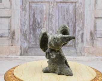 May include: A green stone sculpture of a bird with its wings spread, perched on a rock. The sculpture is sitting on a round, gold-painted table with intricate carvings.