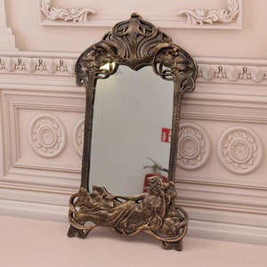 May include: An ornate gold-toned mirror with a decorative frame featuring a reclining figure and floral motifs. The mirror is freestanding and has a rectangular shape.