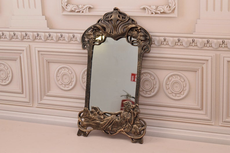 May include: An ornate gold-toned mirror with a decorative frame featuring a reclining figure and floral motifs. The mirror is freestanding and has a rectangular shape.
