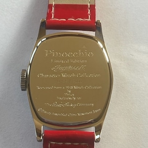 May include: Back of a silver watch with a red leather band. The watch face is engraved with the text "Pinocchio Limited Edition Ingersoll Character Watch Collection Disney 1928", "Licensed from a 1928 Watch Collection by Ingersoll Exclusively for The Walt Disney Company Disney Assembled China Movement Japan".