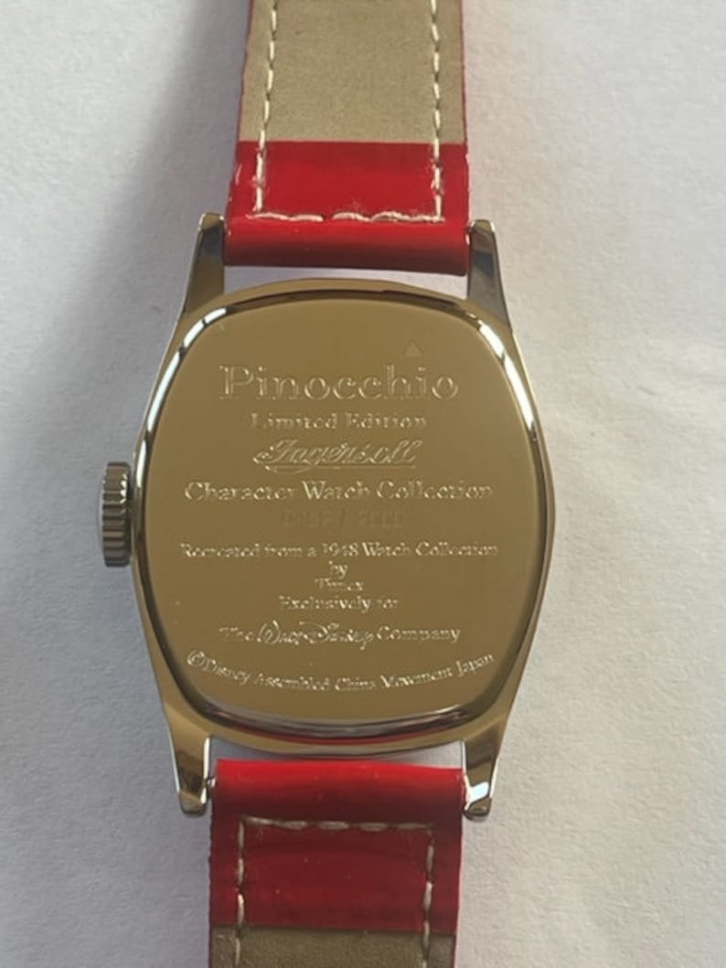 May include: Back of a silver watch with a red leather band. The watch face is engraved with the text "Pinocchio Limited Edition Ingersoll Character Watch Collection Disney 1928", "Licensed from a 1928 Watch Collection by Ingersoll Exclusively for The Walt Disney Company Disney Assembled China Movement Japan".