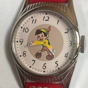May include: A silver wristwatch with a red leather band. The watch face features a cartoon image of Pinocchio with a white background and black Roman numeral hour markers.