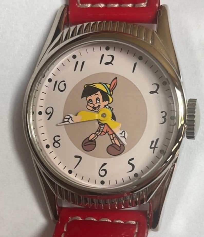 May include: A silver wristwatch with a red leather band. The watch face features a cartoon image of Pinocchio with a white background and black Roman numeral hour markers.