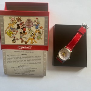 May include: A red box with a Disney character illustration on the front and a certificate of authenticity for a Pinocchio watch. The watch has a red band and a white face with a picture of Pinocchio on it. The watch is in a black box.