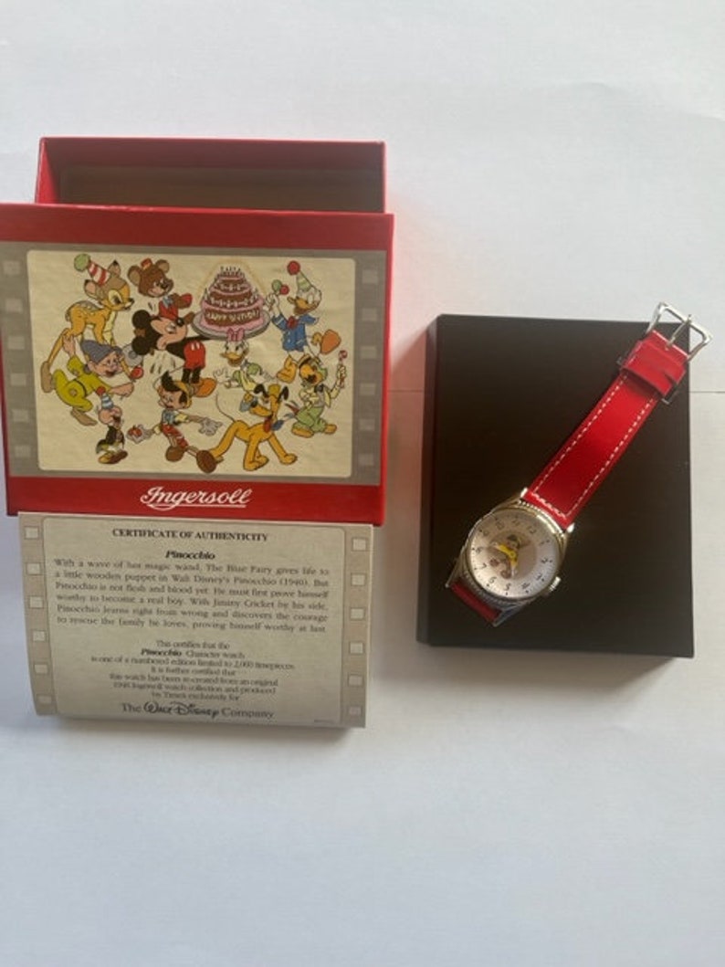 May include: A red box with a Disney character illustration on the front and a certificate of authenticity for a Pinocchio watch. The watch has a red band and a white face with a picture of Pinocchio on it. The watch is in a black box.