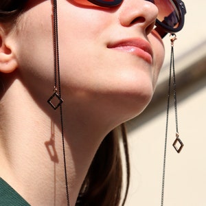 Puede incluir: Una mujer lleva gafas de sol con una cadena negra unida a las monturas. La cadena tiene abalorios cuadrados en los extremos.