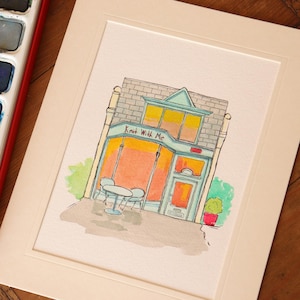 May include: A watercolor illustration of a storefront with the text Knit With Me on the facade. The building features a teal frame, orange-tinted windows, and a small outdoor table with two chairs. The artwork is presented on a white mat board.