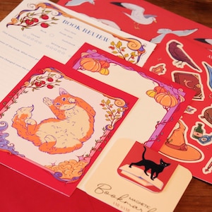 Könnte beinhalten: Ein Schreibwaren-Set bestehend aus einem Buchrezensionsblatt, einer quadratischen Karte mit einer orangefarbenen Katzenillustration, einem roten Stickerbogen mit Rabe, Kessel und Hexenhut sowie einem magnetischen Lesezeichen.